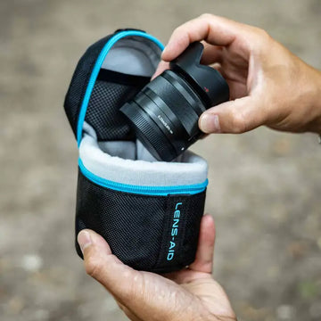 Schwarzes Kameralinsenobjektiv mit griffiger, strukturierter Oberfläche und blauem Ziernähten, eingebettet in eine schwarze Mesh-Tragetasche mit der Aufschrift „lens-aid“