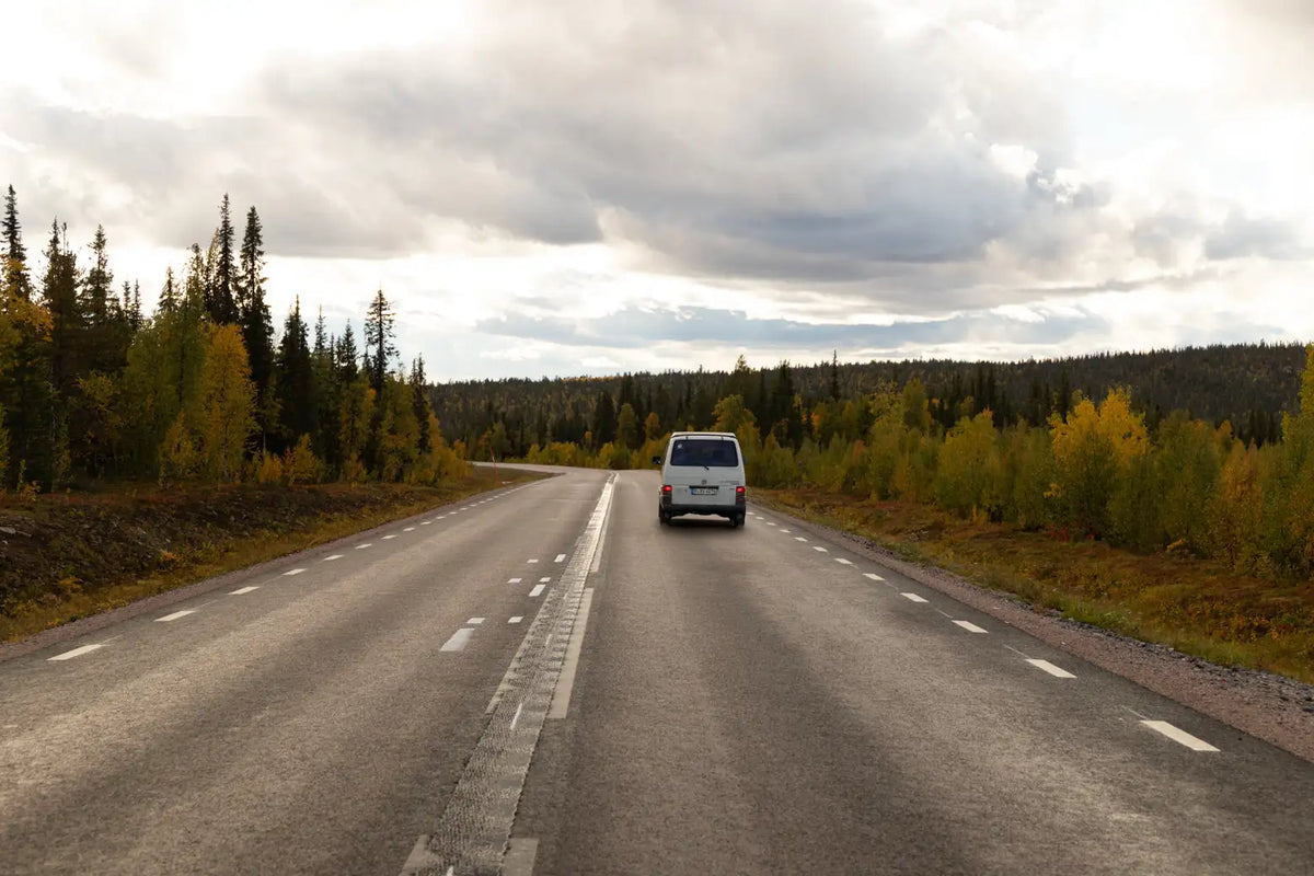 Ein weißer Transporter fährt eine kurvenreiche Straße entlang, gesäumt von Bäumen in Herbstfarben unter einem wolkenverhangenen Himmel.